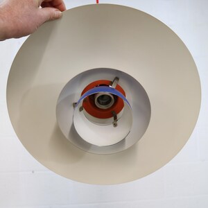 Puede incluir: Una l&aacute;mpara colgante en capas de color crema. La luz tiene un dise&ntilde;o circular con una serie de anillos conc&eacute;ntricos. El centro presenta detalles en naranja y azul. La l&aacute;mpara est&aacute; siendo sostenida por una mano.