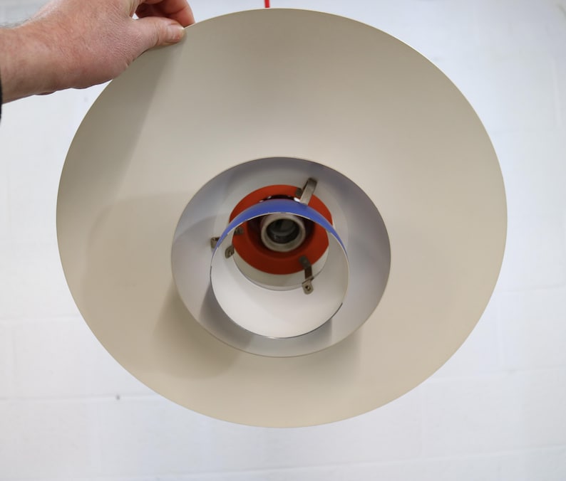 Puede incluir: Una l&aacute;mpara colgante en capas de color crema. La luz tiene un dise&ntilde;o circular con una serie de anillos conc&eacute;ntricos. El centro presenta detalles en naranja y azul. La l&aacute;mpara est&aacute; siendo sostenida por una mano.