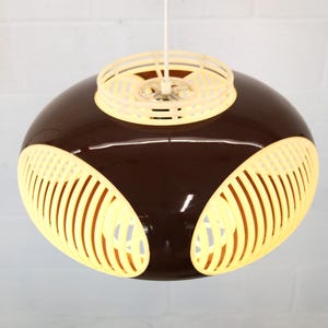 May include: A mid-century modern pendant light with a brown, rounded body and cream-colored accents. The light has a circular top and three sections of vertical slats, creating a unique design. The light hangs from a white cord.