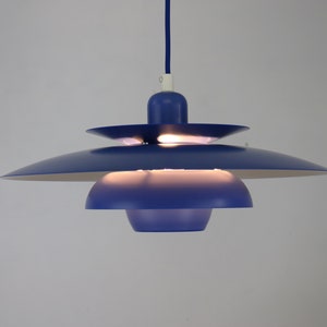 Op de afbeelding: Een blauwe hanglamp met meerdere lagen ronde lampenkappen. De lamp is aan en verlicht de kamer.