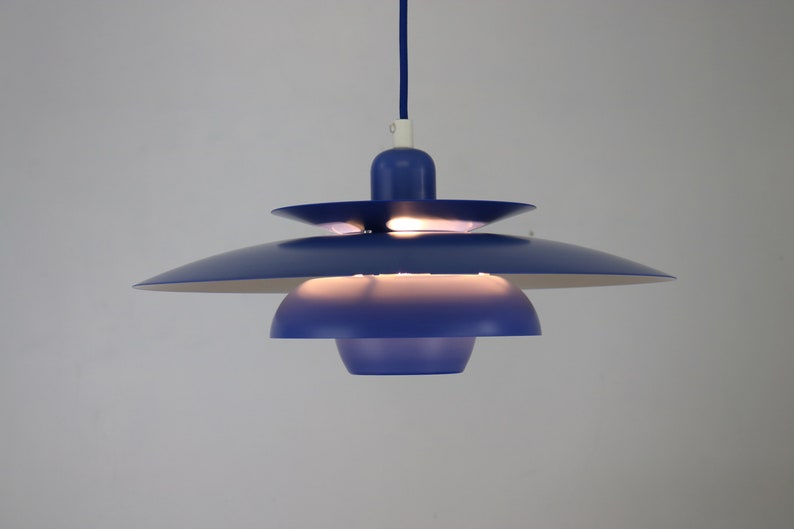 Op de afbeelding: Een blauwe hanglamp met meerdere lagen ronde lampenkappen. De lamp is aan en verlicht de kamer.