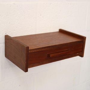Op de afbeelding: Een aan de muur gemonteerde zwevende plank van middelbruin hout met een rechthoekig bovenoppervlak en een enkele geïntegreerde lade met een horizontale houten handgreep in het midden van het voorpaneel voor compacte opslag.