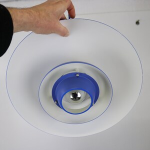 Op de afbeelding: Een witte en blauwe plafondlamp met een cirkelvormig ontwerp. De lamp heeft een blauw midden met een witte ring eromheen. De lamp hangt aan het plafond.