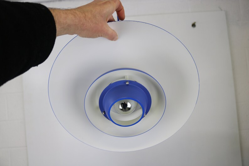 Op de afbeelding: Een witte en blauwe plafondlamp met een cirkelvormig ontwerp. De lamp heeft een blauw midden met een witte ring eromheen. De lamp hangt aan het plafond.