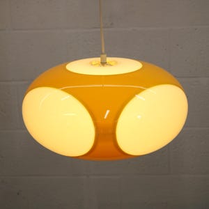 May include: A retro-style pendant light fixture. The light has a rounded, orb-like shape with a mustard yellow exterior and three large, oval-shaped white light panels. The fixture hangs from a thin, white cord.