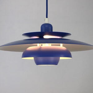 Op de afbeelding: Een blauwe hanglamp met meerdere lagen ronde lampenkappen. De lamp is aan en verlicht de kamer.
