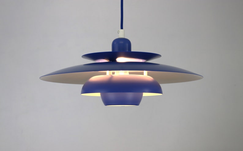Op de afbeelding: Een blauwe hanglamp met meerdere lagen ronde lampenkappen. De lamp is aan en verlicht de kamer.