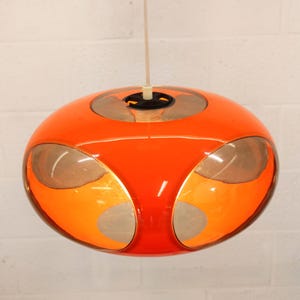 May include: A retro-style orange pendant light fixture. The light has a rounded, orb-like shape with silver and orange accents. It hangs from a white cord, and is suspended against a white brick wall.
