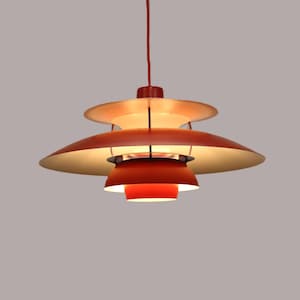 May include: A multi-tiered pendant light fixture featuring a series of stacked, circular metal shades in varying shades of red and orange. The lamp is suspended by a thin red cord and emits a warm, diffused glow from its central light source.