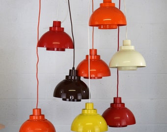 May include: A collection of pendant lights in various colors, including red, orange, yellow, brown, and cream. Each light has a rounded, dome-shaped shade and a cylindrical top, suspended by a cord. The lights are arranged at different heights.
