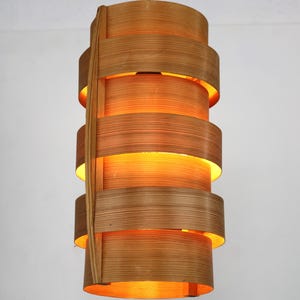 May include: A cylindrical wooden pendant light with a warm, orange glow. The light is made from thin strips of wood that are layered and woven together to create a unique and textured design.
