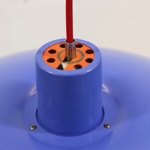 Puede incluir: Una moderna l&aacute;mpara colgante con una pantalla azul y una carcasa cil&iacute;ndrica azul. La l&aacute;mpara tiene un interior naranja y un cable de alimentaci&oacute;n rojo. La luz est&aacute; suspendida del techo.