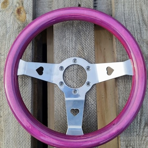Puede incluir: Un volante personalizado con un borde de madera rosa vibrante y un centro de metal plateado pulido. Los radios metálicos presentan recortes en forma de corazón. El volante está diseñado para uso automotriz y se encuentra sobre un fondo de madera.