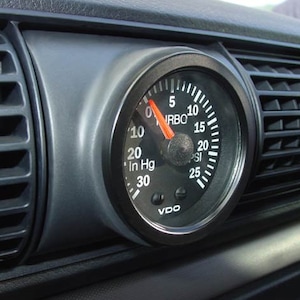 May include: A black VDO boost gauge with a white face and red needle. The gauge reads up to 30 inches of mercury and 25 PSI. The gauge is labeled "Turbo" and has a black bezel.