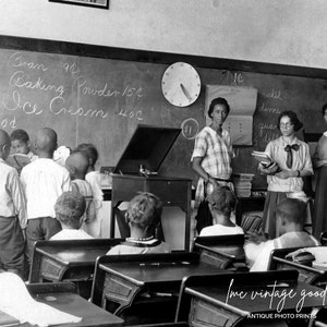 May include: Black and white photo of a classroom with a teacher and students. The teacher is standing at the front of the class, and the students are sitting at their desks. The classroom is decorated with a chalkboard that lists prices for various items, such as pickles, salmon, and ice cream.