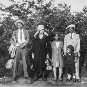 May include: A black and white vintage photograph of a family of five standing in front of a row of trees. The family is dressed in formal attire, including hats and suits. The photo is likely from the 1920s or 1930s.