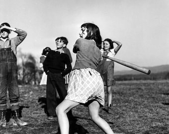 Może przedstawiać: Czarno-białe zdjęcie grupy dzieci grających w baseball. Dziewczynka w wzorzystej sukience macha drewnianą pałką. Zdjęcie zawiera napis "lmc vintage goods" na dole.
