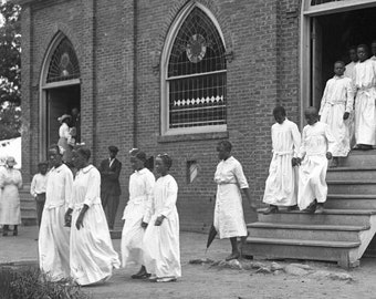 Puede incluir: Una fotografía en blanco y negro de un grupo de personas con túnicas blancas de pie frente a un edificio de ladrillo con una gran puerta de madera. Las personas miran todas a la cámara. El edificio tiene una gran ventana con vidrieras.