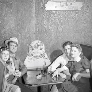 May include: A black and white photo of four people sitting at a table in a bar. The sign above the table reads 
