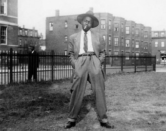 May include: Black and white photo of a man wearing a suit and hat, standing in front of a fence with buildings in the background. He is holding a cigarette in his mouth.