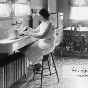 May include: Vintage black and white photograph of a woman in a kitchen. She is seated at a stool, working at a large sink. A pot sits on the sink's edge. The kitchen features a stove, table, and chairs, with a window in the background.