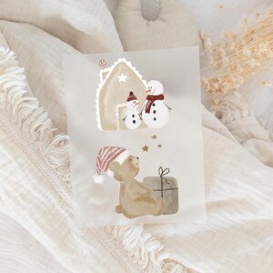 Könnte beinhalten: Eine Grußkarte mit Aquarellillustrationen eines Lebkuchenhauses, Schneemännern, einem Teddybären und einem verpackten Geschenk. Die Schneemänner tragen einen Schal und der Bär trägt eine Wintermütze. Die Karte hat eine weiche, neutrale Farbpalette.