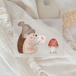 Könnte beinhalten: Aquarellillustration eines Igels mit grauem Hut, der eine Gänseblümchen hält, neben einem rot-weiß gepunkteten Pilz sitzt. Das Kunstwerk hat einen weichen, skurrilen Stil, ideal für Kinderzimmerdekoration.