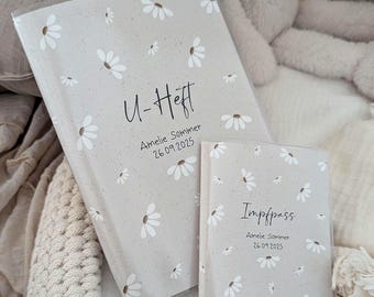 Könnte beinhalten: Zwei hellgraue Bücher mit weißen Gänseblümchenmustern. Das größere Buch trägt den Text "U-Heft" und das kleinere Buch den Text "Impfpass". Beide Bücher tragen den Namen "Amelie Sommer" und das Datum "26.09.2025".