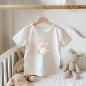 May include: A white short-sleeved t-shirt featuring a graphic print of a large, muted pink number three, a small white duck, a bee, and white daisy flowers. The shirt is displayed on a wooden hanger, showcasing a crew neckline and a simple cut.