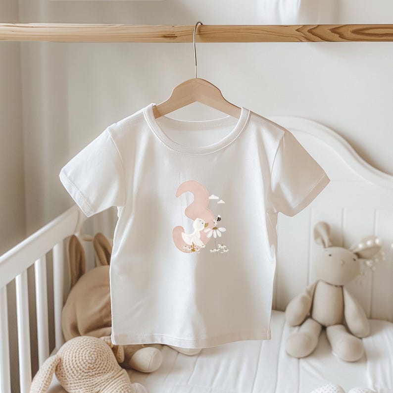 May include: A white short-sleeved t-shirt featuring a graphic print of a large, muted pink number three, a small white duck, a bee, and white daisy flowers. The shirt is displayed on a wooden hanger, showcasing a crew neckline and a simple cut.