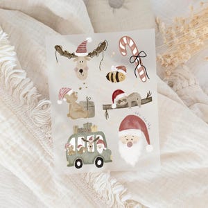 Könnte beinhalten: Aquarellillustrationen mit Weihnachtsmotiven. Enthält ein Rentier mit Weihnachtsmütze, eine Zuckerstange, eine Biene, einen Teddybären, Faultiere, einen grünen Van mit Geschenken und den Weihnachtsmann. Die Illustrationen sind in sanften, gedämpften Farben gehalten.