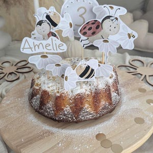 Könnte beinhalten: Ein mit Puderzucker bestäubter Gugelhupf, verziert mit Gänseblümchen- und Insekten-Themen-Toppern. Die Topper zeigen einen Marienkäfer, eine Biene und den Namen "Amelie". Ein runder Topper zeigt die Zahl "3". Der Kuchen steht auf einem Holzbrett mit goldenem Konfetti.