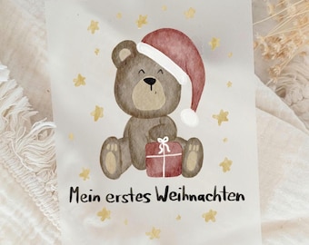 Puede incluir: Tarjeta de Navidad con una ilustración en acuarela de un oso de peluche marrón con un gorro de Papá Noel y un regalo rojo. La tarjeta tiene detalles de estrellas doradas y el texto "Mein erstes Weihnachten" en negro.