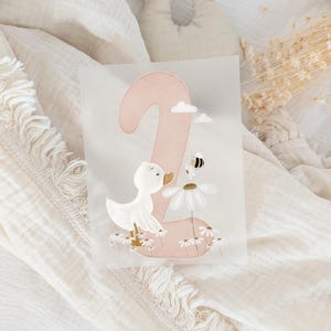 May include: A rectangular card featuring a large, soft pink number two. The design includes a white duckling standing among small white daisies, a larger daisy with a small bumblebee hovering above it, and two tiny white clouds in the upper area.