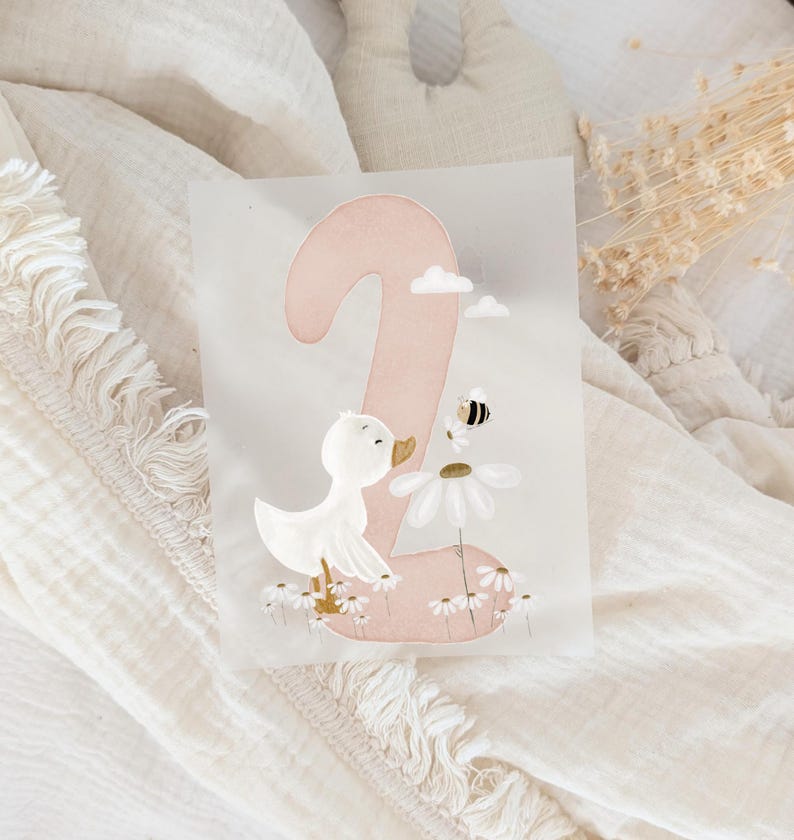 May include: A rectangular card featuring a large, soft pink number two. The design includes a white duckling standing among small white daisies, a larger daisy with a small bumblebee hovering above it, and two tiny white clouds in the upper area.