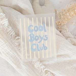 Könnte beinhalten: Eine rechteckige, halbtransparente Karte mit vertikalen blassgelben Streifen und dem Text Cool Boys Club, der in einer hellblauen, abgerundeten Schriftart gedruckt ist. Die Karte liegt auf einem weichen, strukturierten, cremeweißen Stoff.
