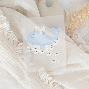 Könnte beinhalten: Eine Illustration eines hellblauen Wals mit einer kleinen weißen Ente auf dem Rücken, umgeben von weißen Gänseblümchen mit goldenen Zentren. Das Kunstwerk befindet sich auf transparentem Papier vor einem weichen, cremefarbenen Hintergrund.