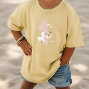 May include: A pale yellow short-sleeved t-shirt featuring a large pink number one graphic. The design includes a white duck, a small bee, and white daisy flowers. The shirt has a crew neckline and a loose, relaxed fit, providing a casual appearance.