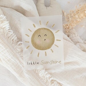 Könnte beinhalten: Aquarell-Illustration einer lächelnden Sonne in Beige- und Brauntönen mit Strahlen auf weißem Hintergrund. Der Text "little Sunshine" steht darunter. Das Bild befindet sich auf einem cremefarbenen, strukturierten Stoff.
