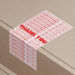 May include: A white sticker with the words "THANK YOU" repeated in a red, stylized font. The sticker is wrapped around the corner of a cardboard box, with the text visible on both sides.