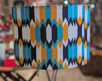 May include: Cylindrical lampshade with a geometric pattern in shades of blue, yellow, black, and white. The fabric shade is mounted on a silver metal lamp base. The pattern features vertical rows of diamond shapes.