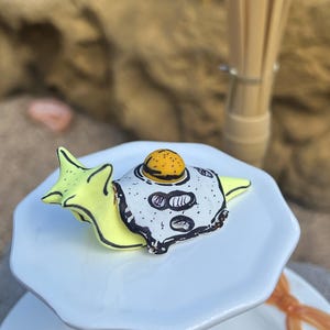 May include: A whimsical, hand-painted ceramic snail figurine with a fried egg shell. The snail is yellow with a white and black eggshell design. The figurine sits on a white, tiered serving plate. The overall theme is food and novelty.