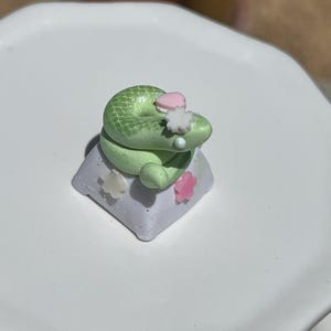 May include: A light green coiled snake figurine sits atop a square, light purple keycap. The snake is decorated with small, white and pink star-shaped accents and a tiny pink heart shape on its back, creating a detailed, miniature sculptural piece.