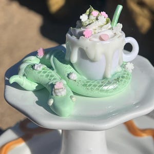 May include: A whimsical sculpture featuring a white teacup overflowing with whipped cream, adorned with pink and white floral accents. The cup rests on a light green snake coiled around a white plate, creating a unique and artistic design.
