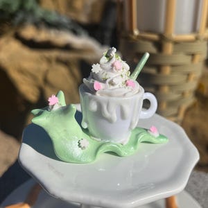 May include: A miniature white teacup with a whipped cream topping, decorated with pink and green accents. The cup sits on a green, leaf-shaped base, placed on a white, tiered stand. A small green spoon rests in the cup.