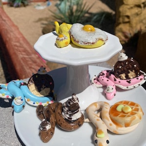 May include: Five miniature ceramic snakes, each with a different food-themed design. The snakes are arranged on a white cake stand and a white plate. The designs include a fried egg, a waffle, a chocolate chip cookie, a donut, and a pancake.