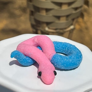 May include: A pink and blue snake-shaped candy with black eyes sits on a white, scalloped-edge plate. The candy is covered in a sugary coating, giving it a sparkling appearance. The background is blurred, with a woven basket visible.