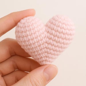 May include: A small, handmade, light pink crochet heart. The heart is held between two fingers, showcasing the textured stitches and soft color. This item is a charming, handcrafted decorative piece.