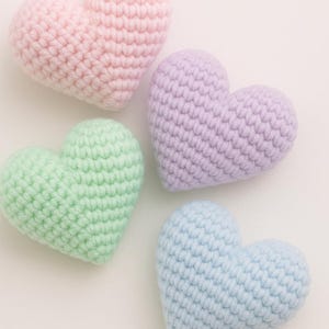 May include: Four handmade crochet hearts in pastel colors: pink, lavender, green, and blue. Each heart is crafted with a textured stitch, creating a soft, plush appearance. The hearts are arranged on a white background, perfect for Valentine's Day or other celebrations.