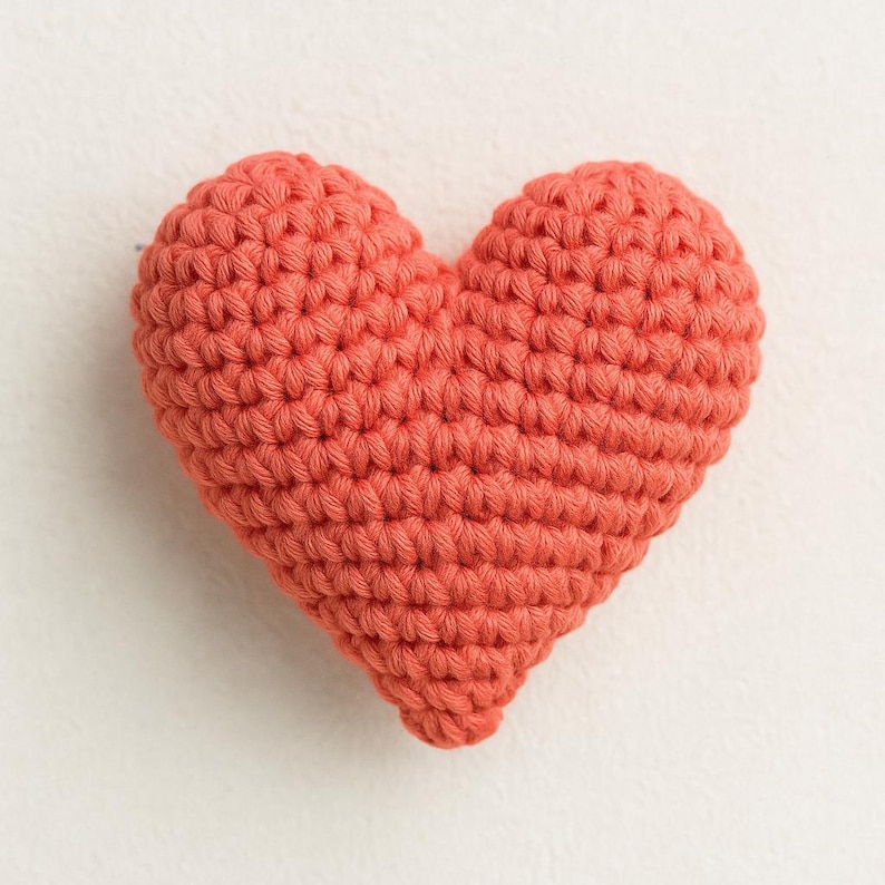 May include: A coral-colored, crocheted heart. The handmade heart is made with a tight stitch pattern, giving it a textured appearance. The heart shape is symmetrical, with a rounded top and a pointed bottom. The craft item is likely a decorative piece or a small toy.